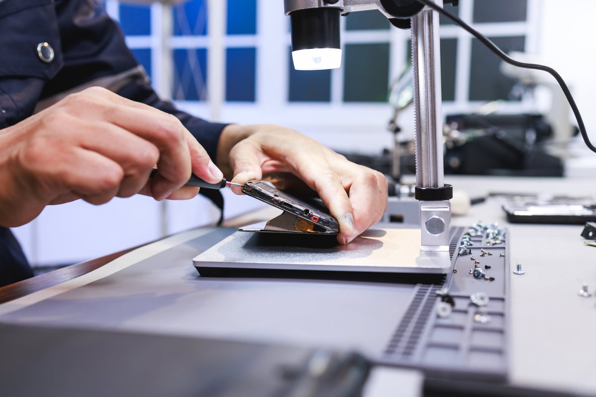 Close-up of a technician repairing a smartphone on a workbench under a small lamp, showing phone maintenance, electronics service, and careful hands-on work in a modern workshop.